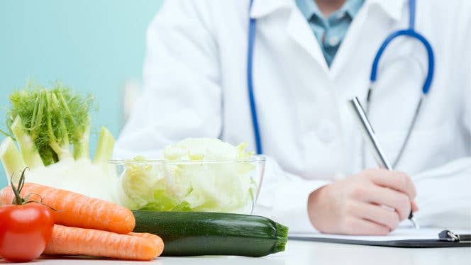 lifestyle medicine A doctor with a white coat and stethoscope writes notes with vegetables on the table in the foreground